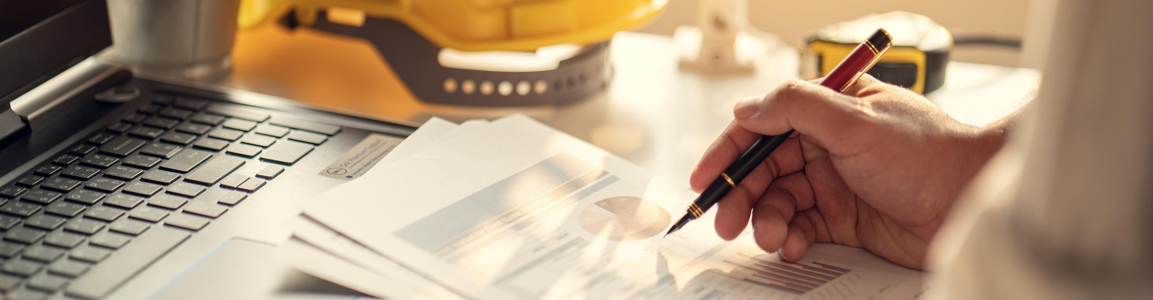Hand holding a pen over construction project charts beside a yellow hard hat and measuring tape