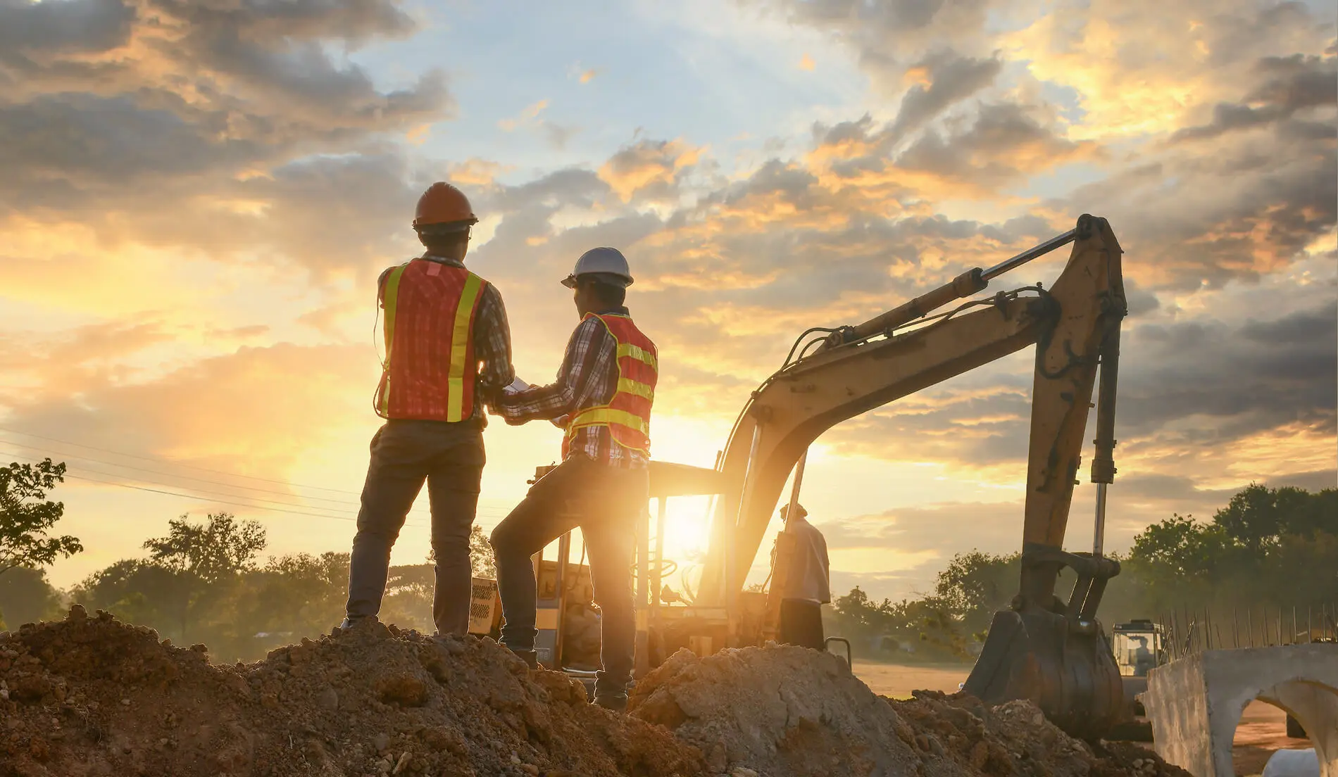Two construction workers in safety vests and hard hats overlooking a job site at sunset beside an excavator