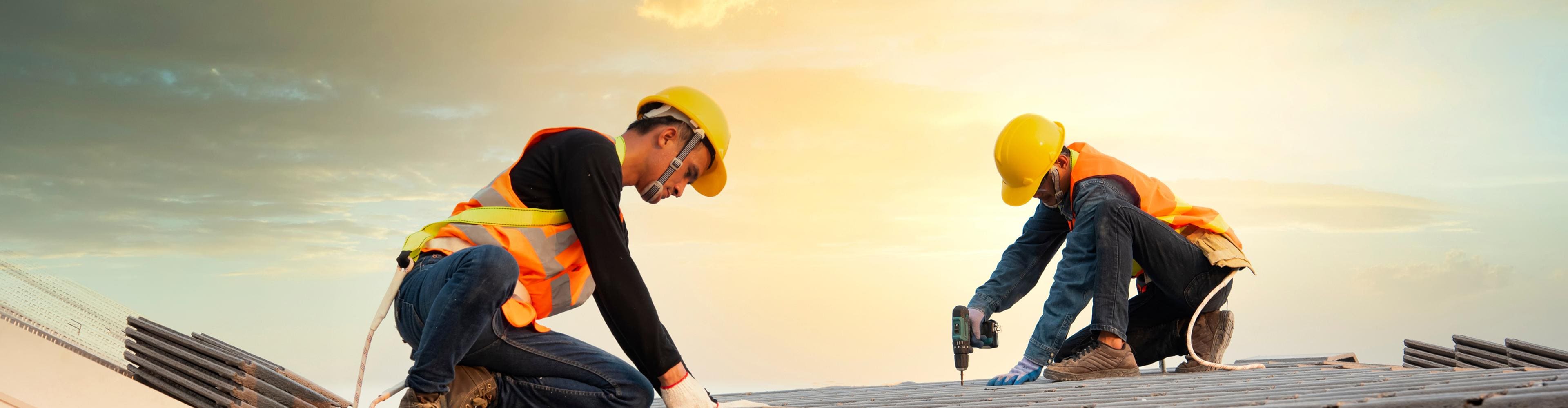 Two roofers in yellow hard hats and orange safety vests installing dark grey roof tiles at sunset