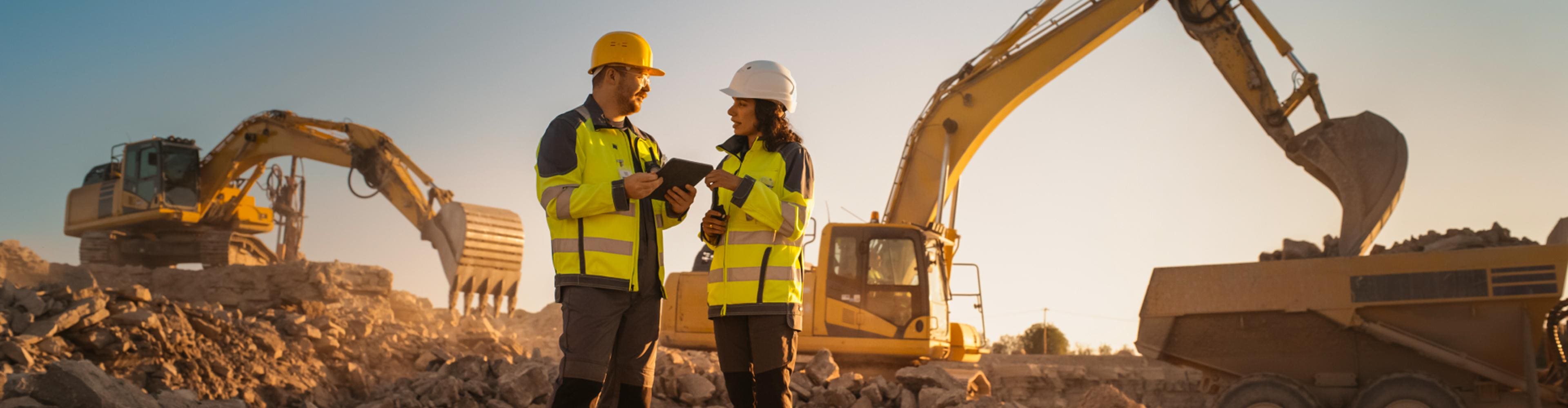 Two construction managers in hard hats discussing plans beside excavators and a dump truck at sunset