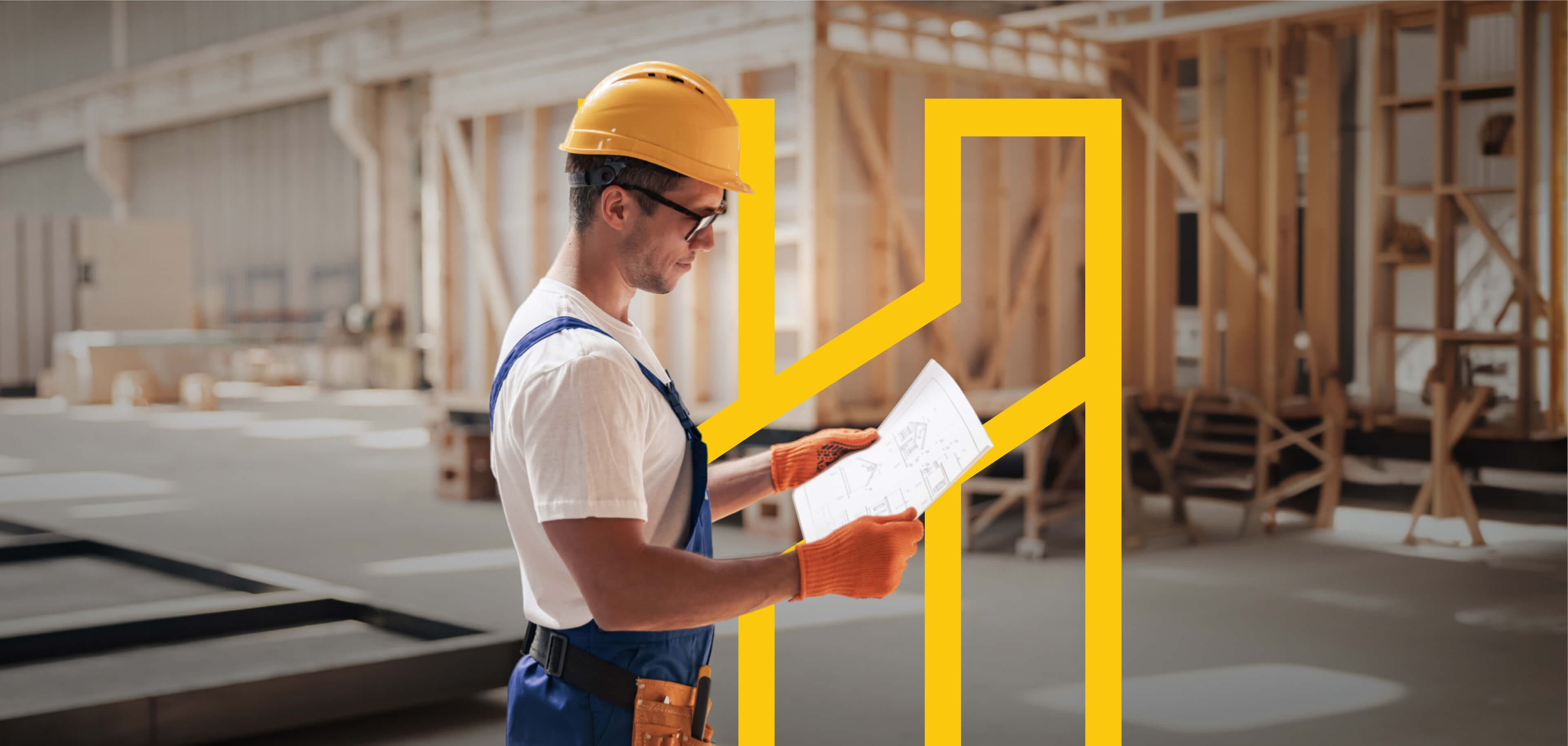 Carpenter in a yellow hard hat and orange gloves studying a house blueprint in a workshop
