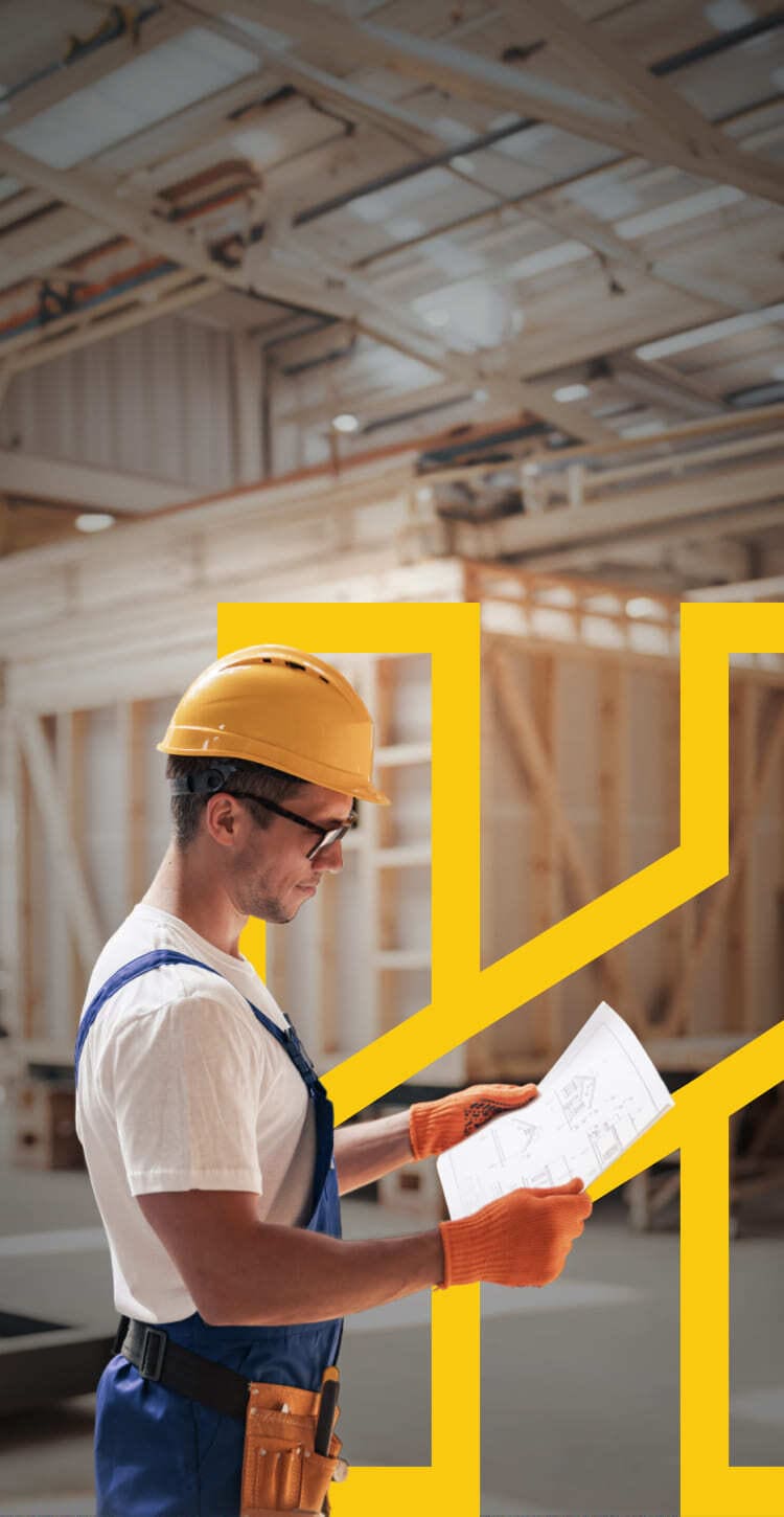 Carpenter in a yellow hard hat and orange gloves studying a house blueprint in a workshop