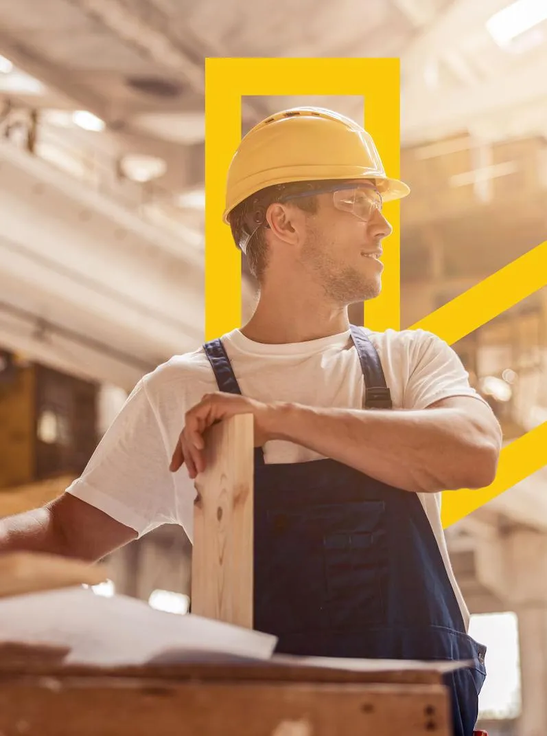 Carpenter in a yellow hard hat, safety glasses, and blue overalls standing by lumber in a workshop