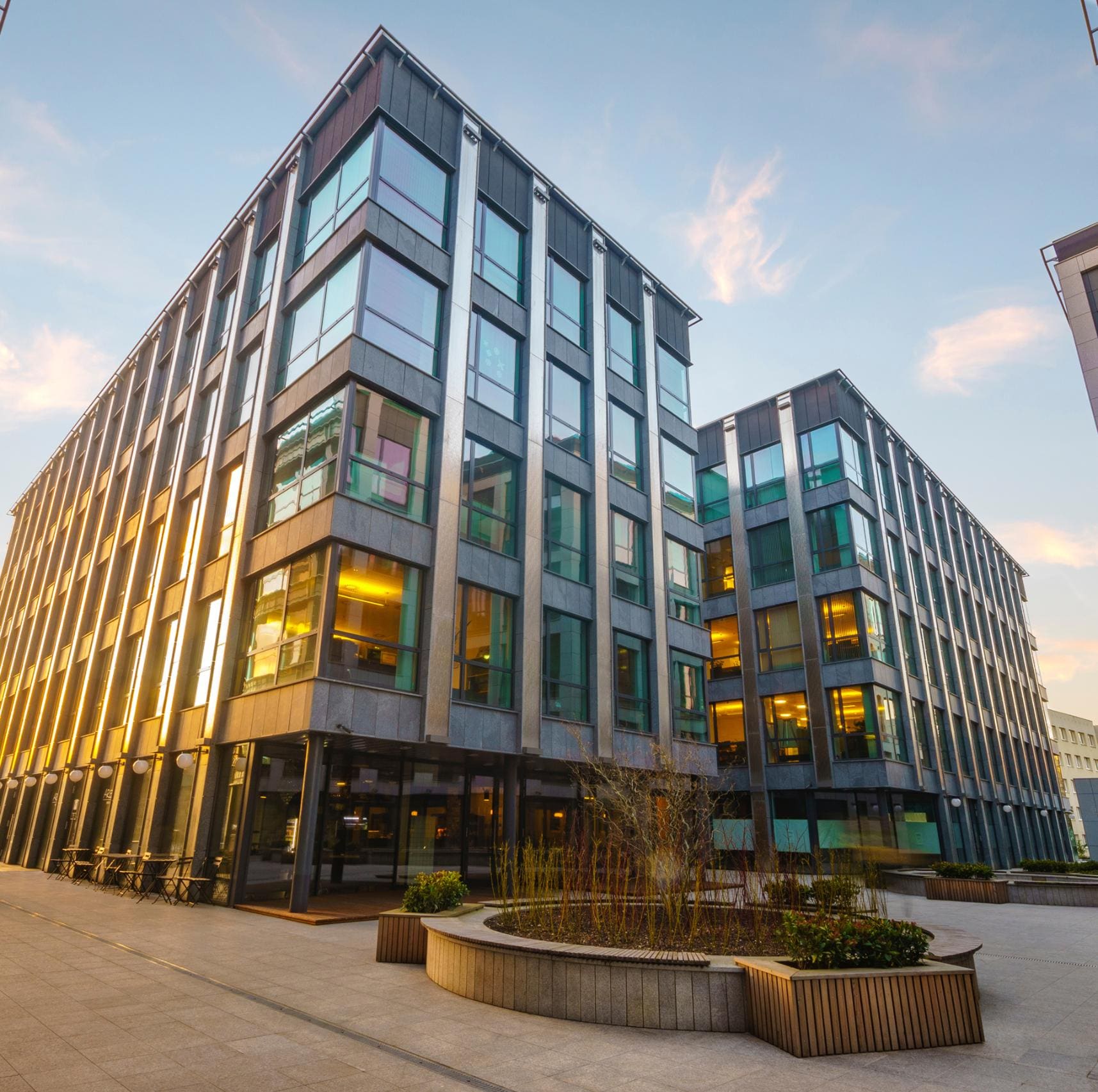 Modern multi-story glass office building at dusk above a paved plaza with planters