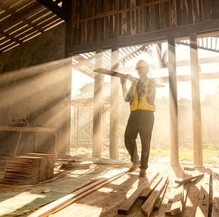 Construction worker carrying a wooden plank through a building frame lit by sunbeams