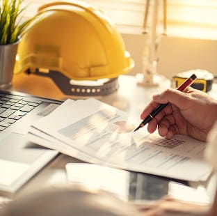 Hand reviewing construction charts on a desk beside a yellow hard hat