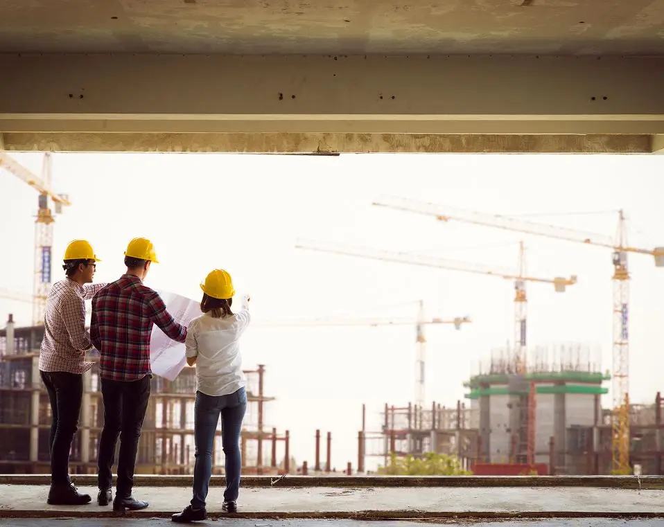 Three construction professionals in yellow hard hats reviewing blueprints overlooking a crane-filled job site