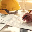 Hand reviewing construction project charts beside a yellow hard hat on a desk