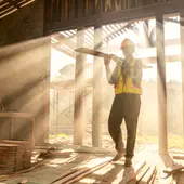 Construction worker in a hard hat and safety vest carrying lumber through a sunlit building frame