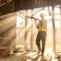 Construction worker in a hard hat and safety vest carrying lumber through a sunlit building frame