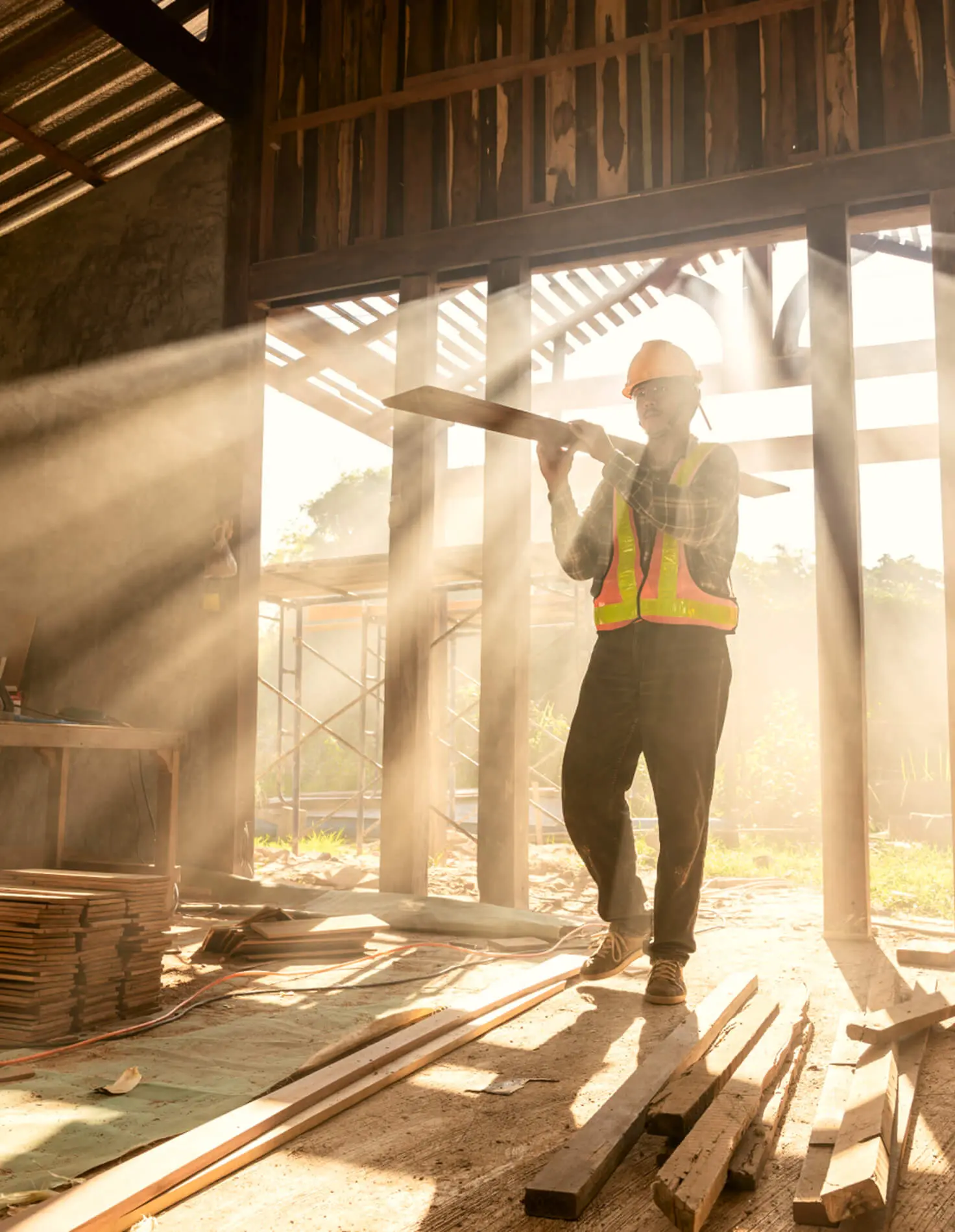 Construction worker in a hard hat and safety vest carrying lumber through a sunlit building frame