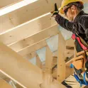 Carpenter in a yellow hard hat and safety harness working among wooden roof rafters