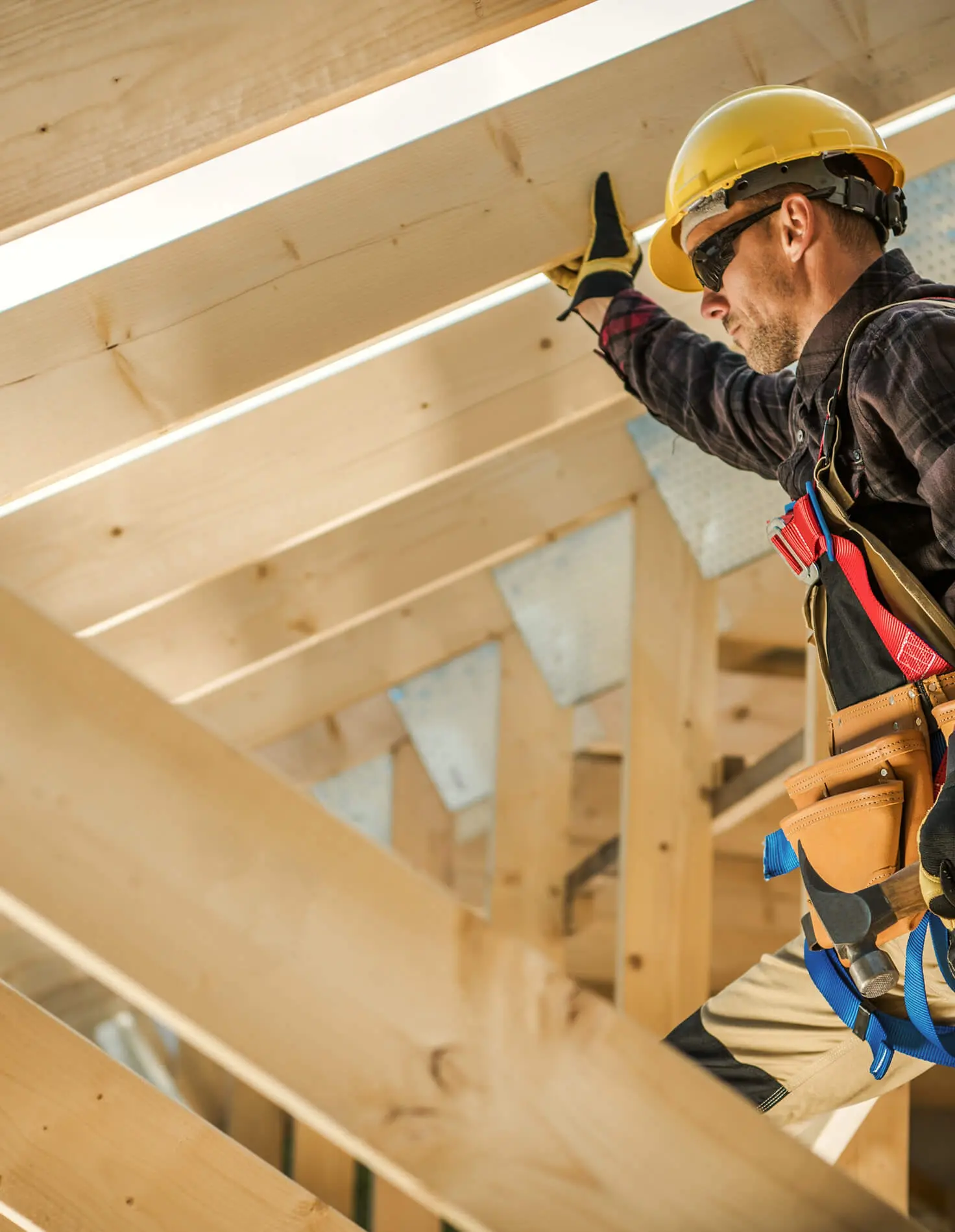 Carpenter in a yellow hard hat and safety harness working among wooden roof rafters