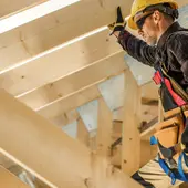 Carpenter in a yellow hard hat and safety harness working among wooden roof rafters