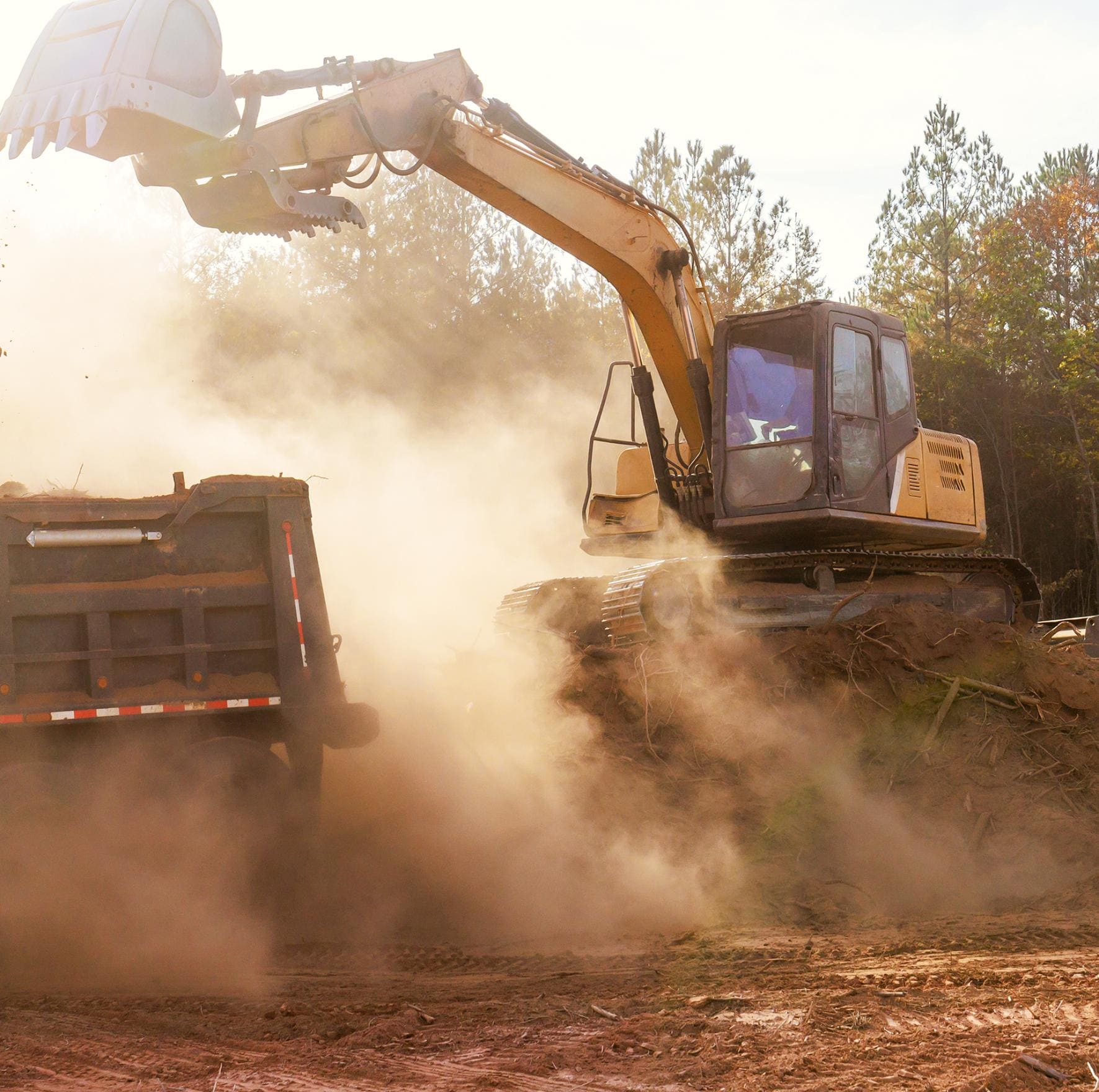 Yellow excavator clearing earth beside a dump truck with dust rising on a job site