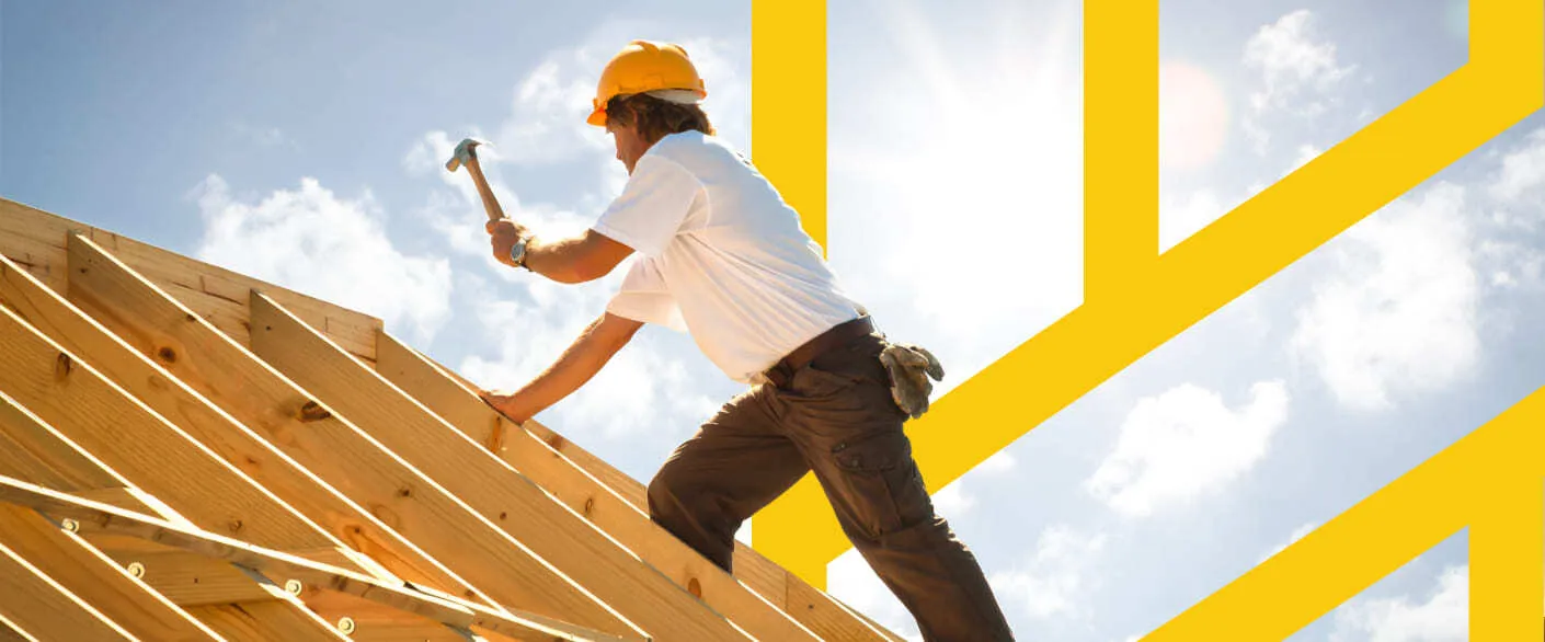 Construction worker in a yellow hard hat hammering wooden roof rafters under a clear blue sky
