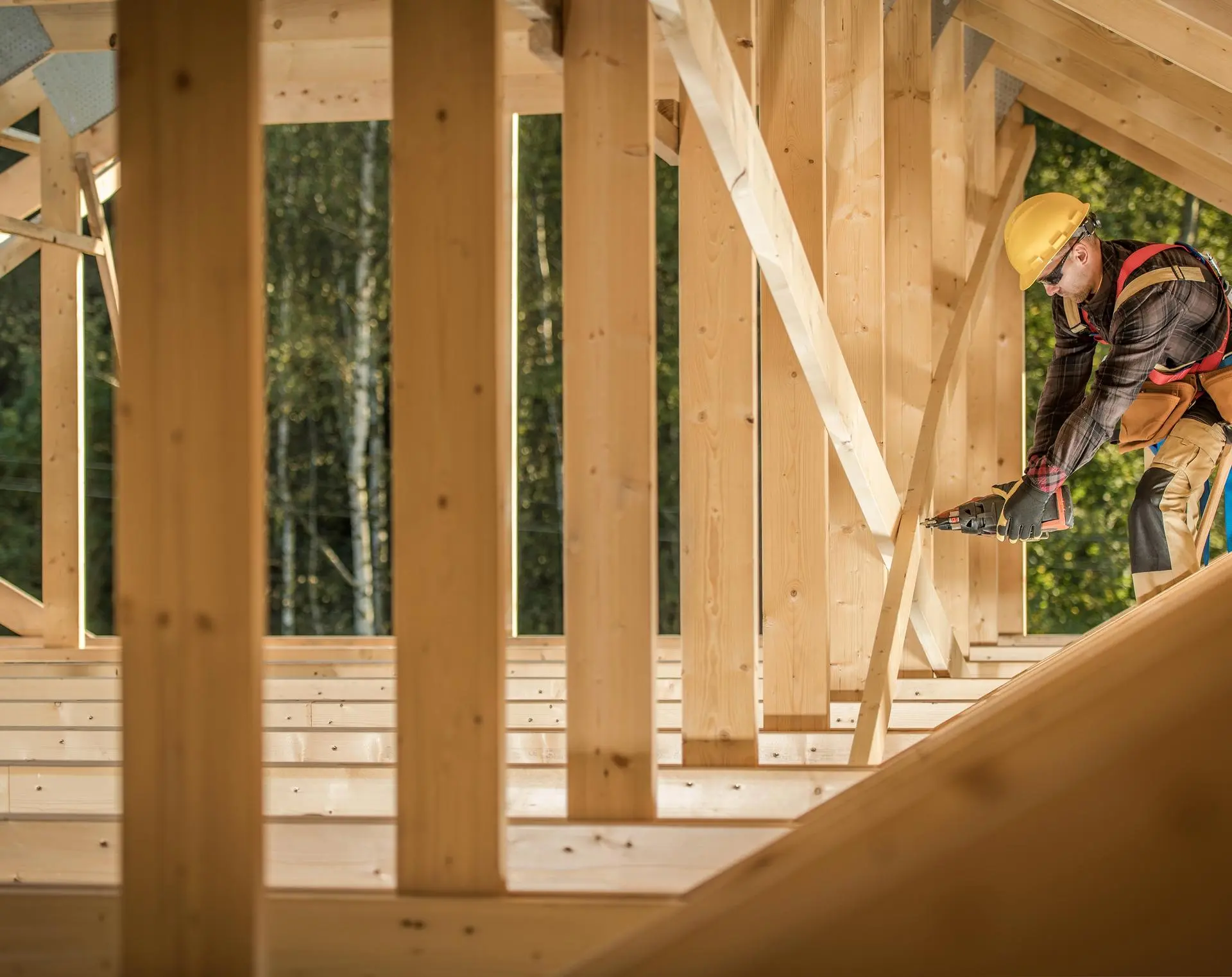 Carpenter in a yellow hard hat and safety harness nailing wooden framing on a new home