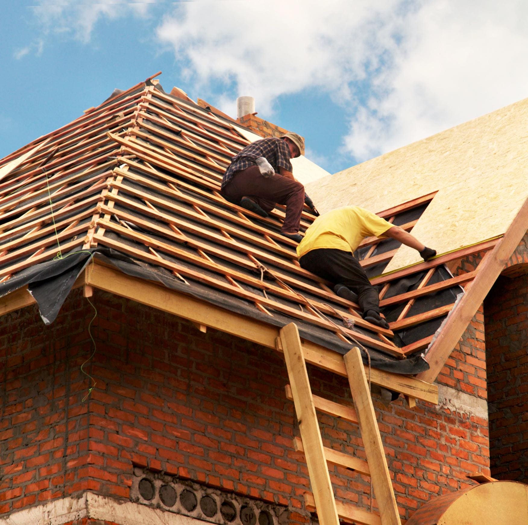 Two workers installing plywood sheathing on a red brick house roof under a blue sky