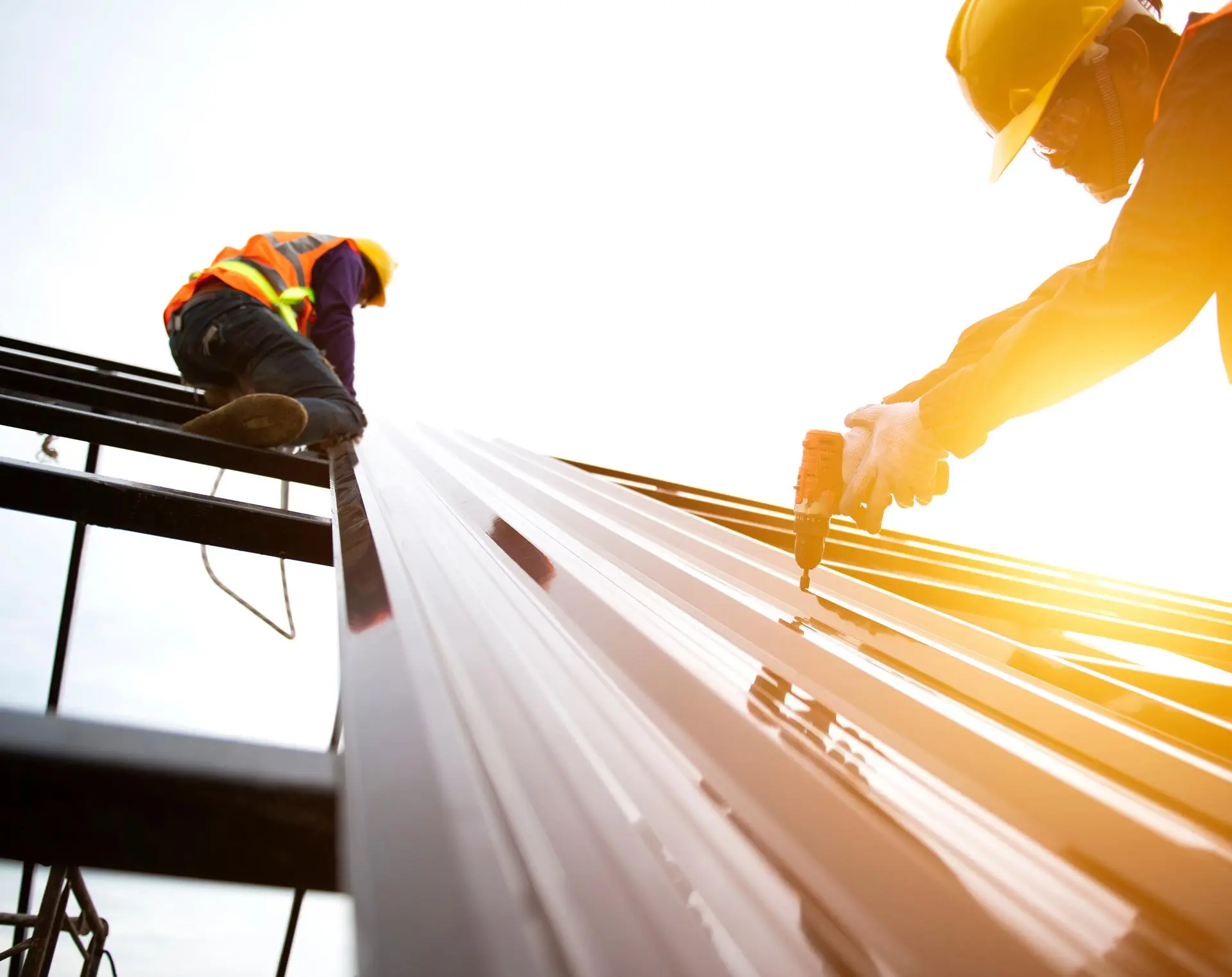 Roofer in a yellow hard hat drilling screws into corrugated metal roof panels in bright sunlight