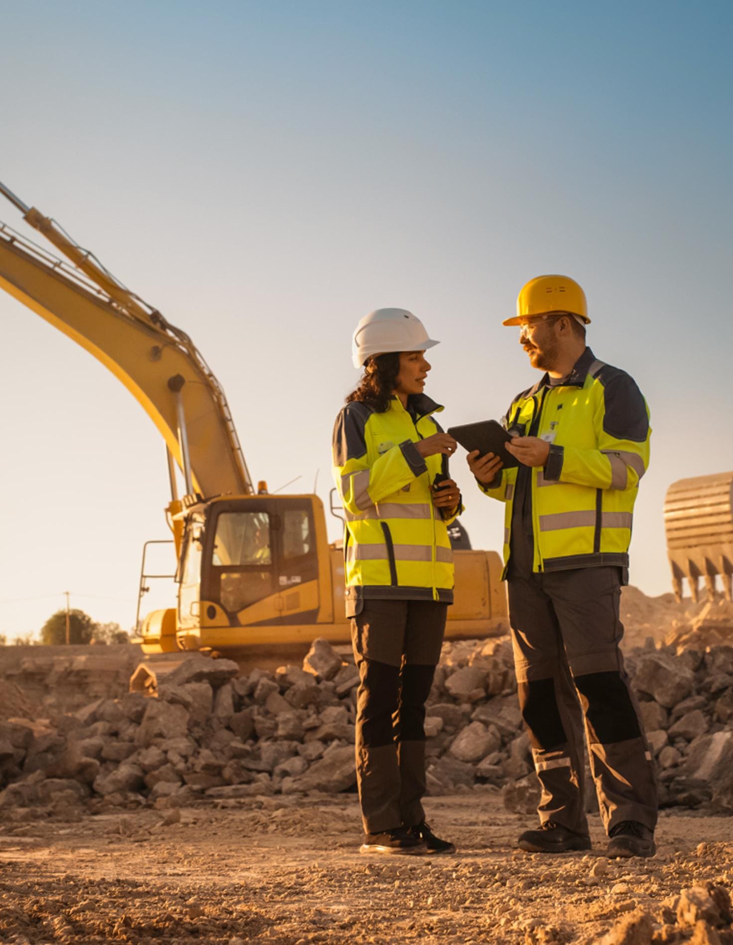 Two site managers in hard hats reviewing a tablet beside a yellow excavator at sunset