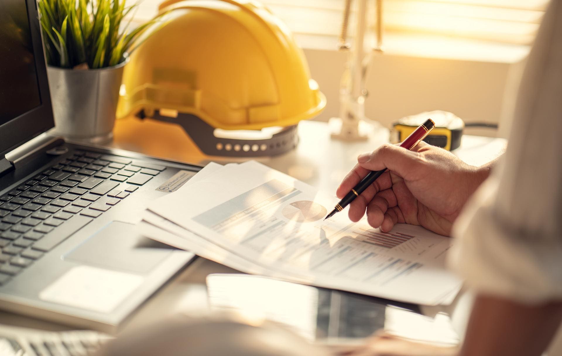 Hand reviewing construction project charts beside a yellow hard hat on a desk