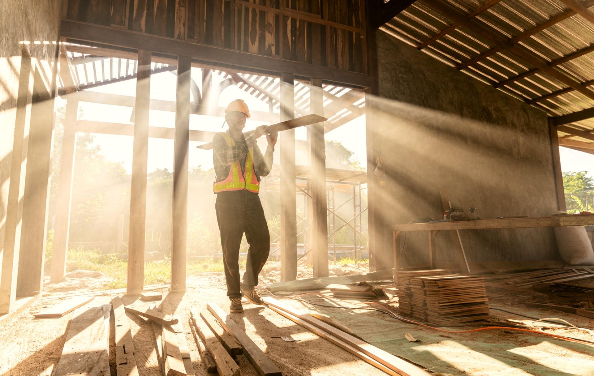 Construction worker in a hard hat and safety vest carrying lumber through a sunlit building frame