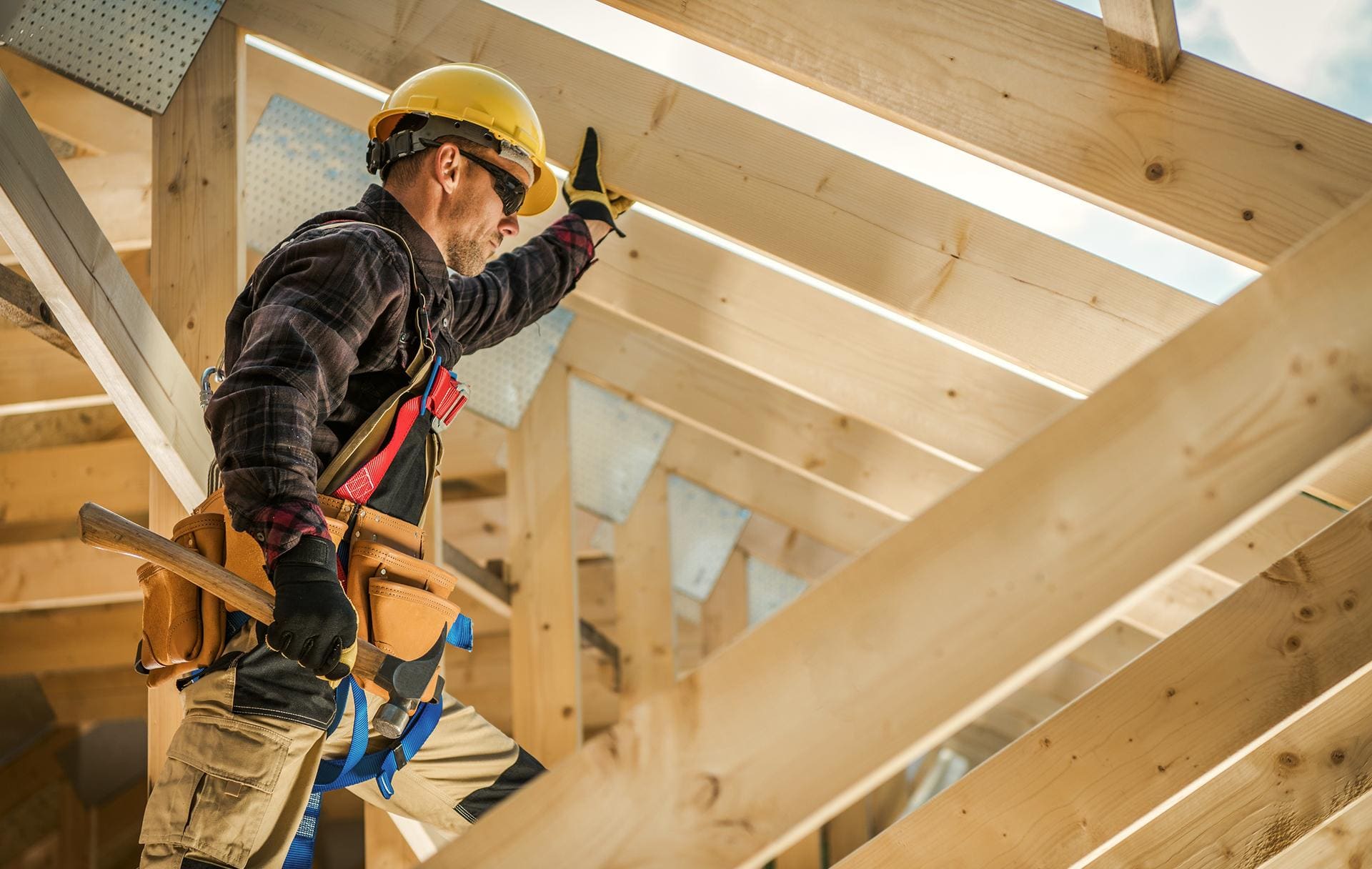 Carpenter in a yellow hard hat and safety harness working among wooden roof rafters