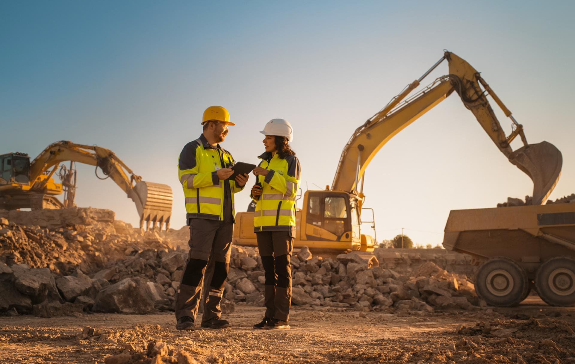 Two site managers in hard hats reviewing a tablet beside excavators at sunset